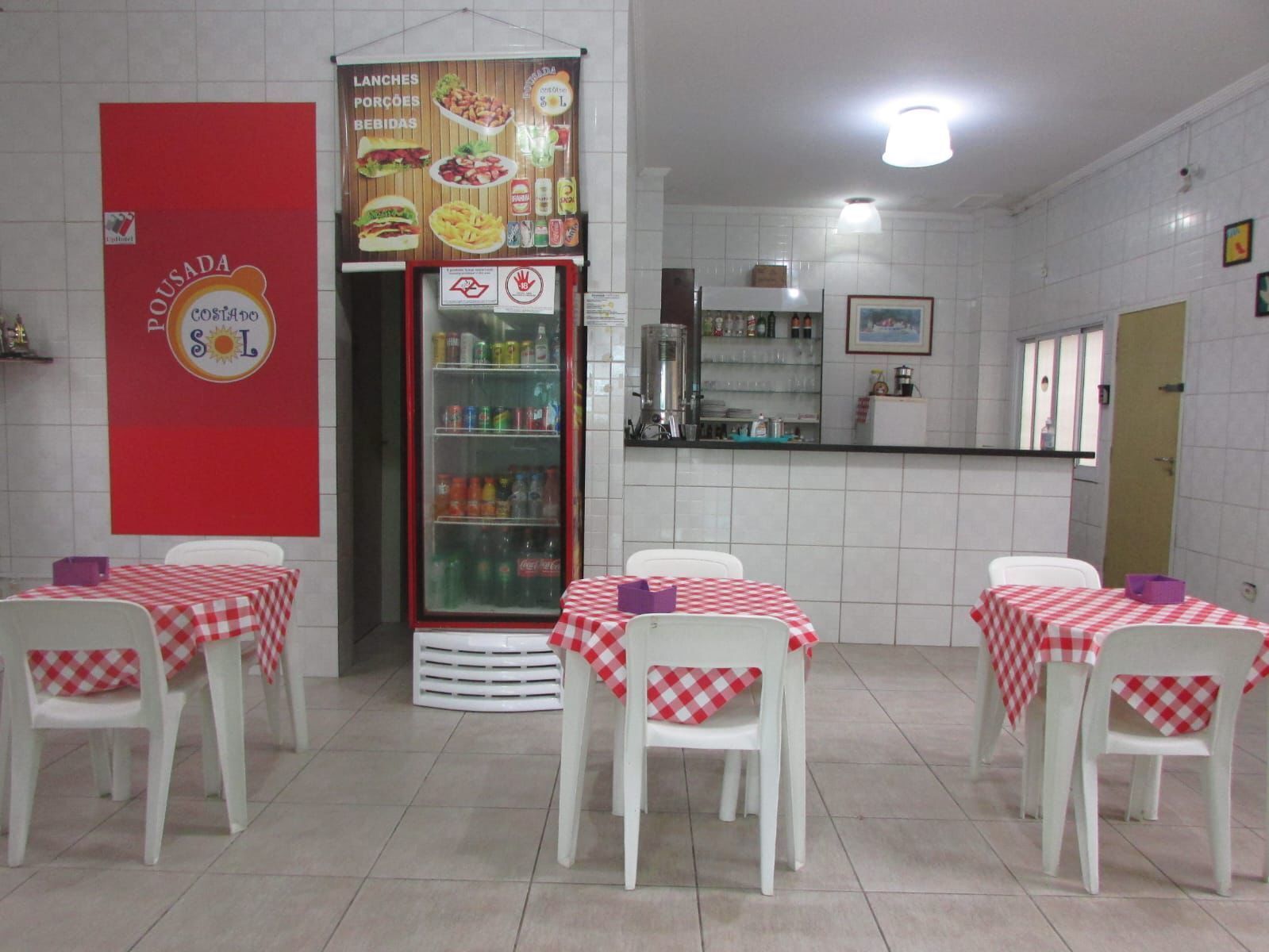 A restaurant with checkered tablecloths, white chairs, a refrigerator stocked with beverages, and a menu board advertising lunches, porcoes, and bebidas.