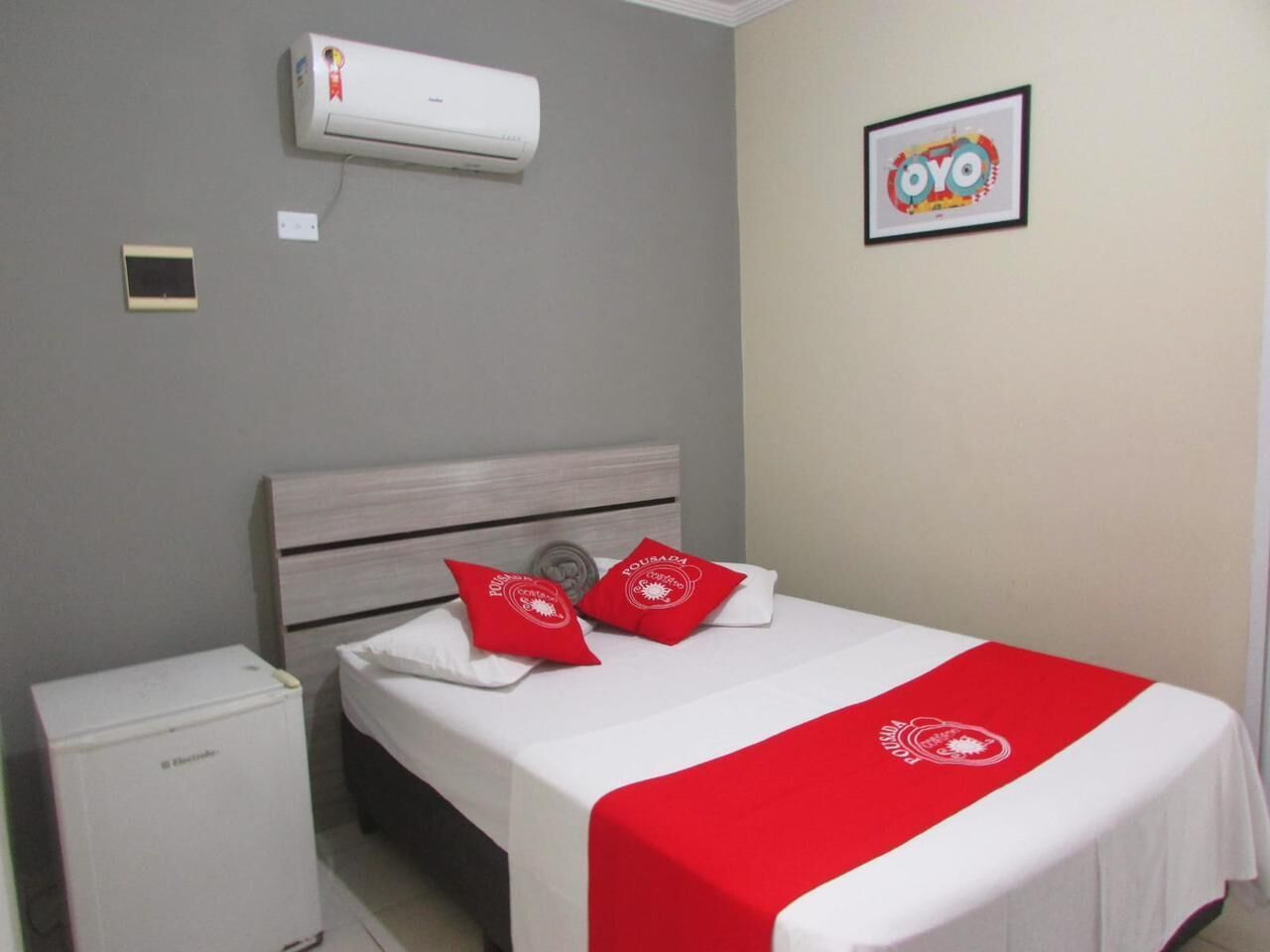 A hotel room with a bed, a small bedside table, an air conditioner, and a framed picture on the wall.