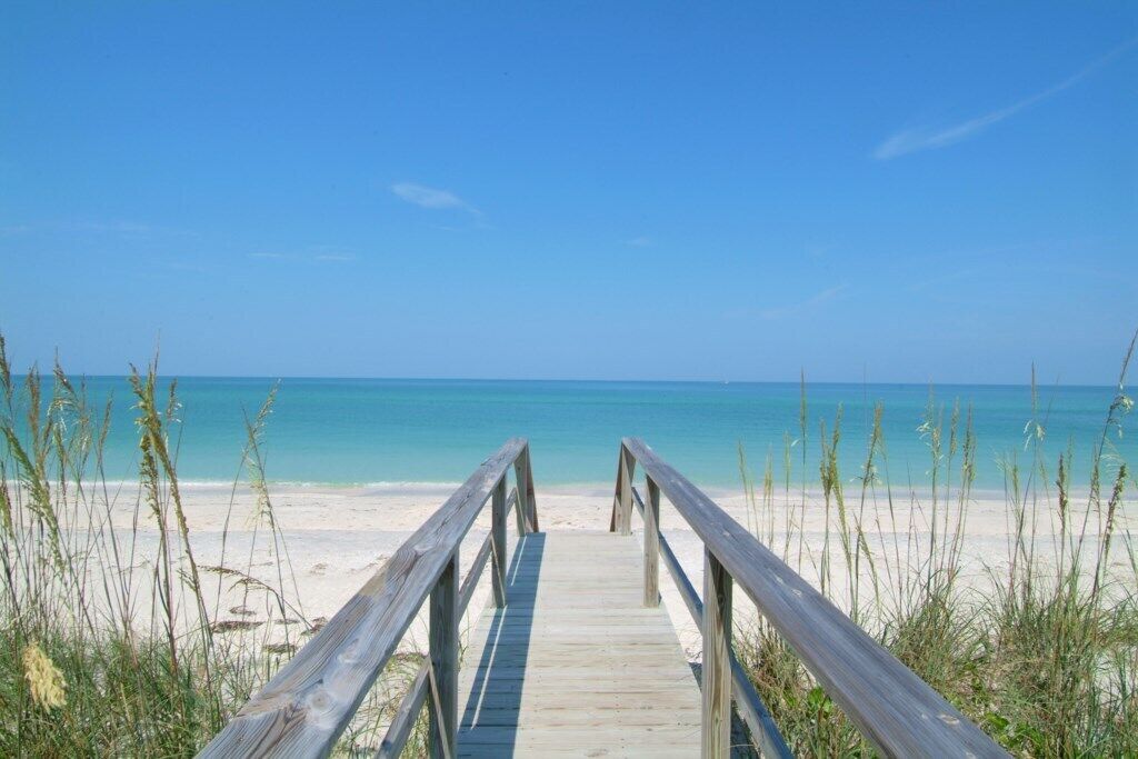 Een houten looppad dat naar een strand leidt met helderblauw water en een zandstrand.