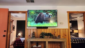 A living room with a television mounted on the wall, a wooden console table with decorative items, and a bed with a checkered blanket. - Rancho Relaxo, large fully fenced pet friendly lot. Centrally located (Big Bear)