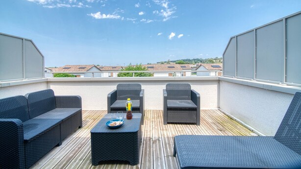 A rooftop terrace with wicker furniture, a wooden deck, and a view of buildings in the distance. - The Cocoon - Appt avec terrasse, 20mn de Toulouse (Castelginest)