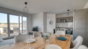A modern dining area with a wooden table set for four, a kitchen in the background, and large windows offering a view of the outdoors. - The Cocoon - Appt avec terrasse, 20mn de Toulouse (Castelginest)