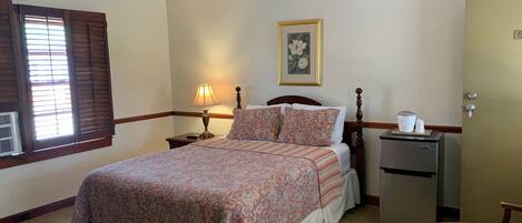 A hotel room with a bed, a bedside table with a lamp, a refrigerator, a door, and a window with blinds.
