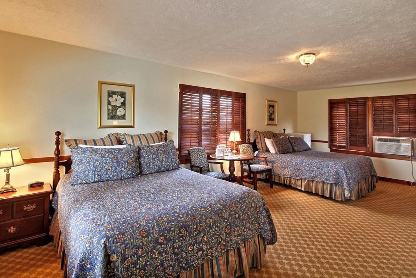 A hotel room with two beds, a desk with a lamp, and a chair.