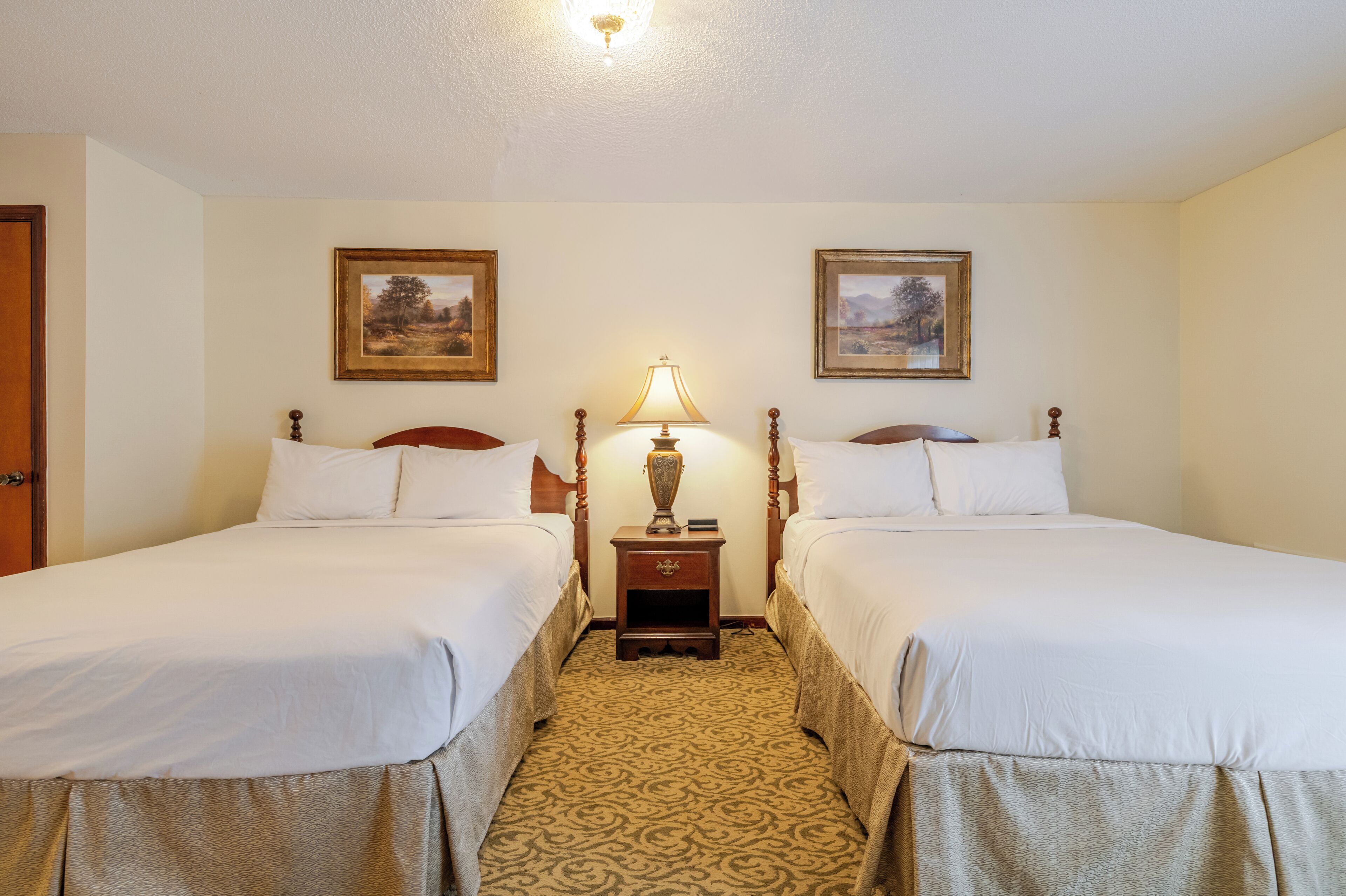 A hotel room with two beds, a nightstand with a lamp, and two framed pictures on the wall.