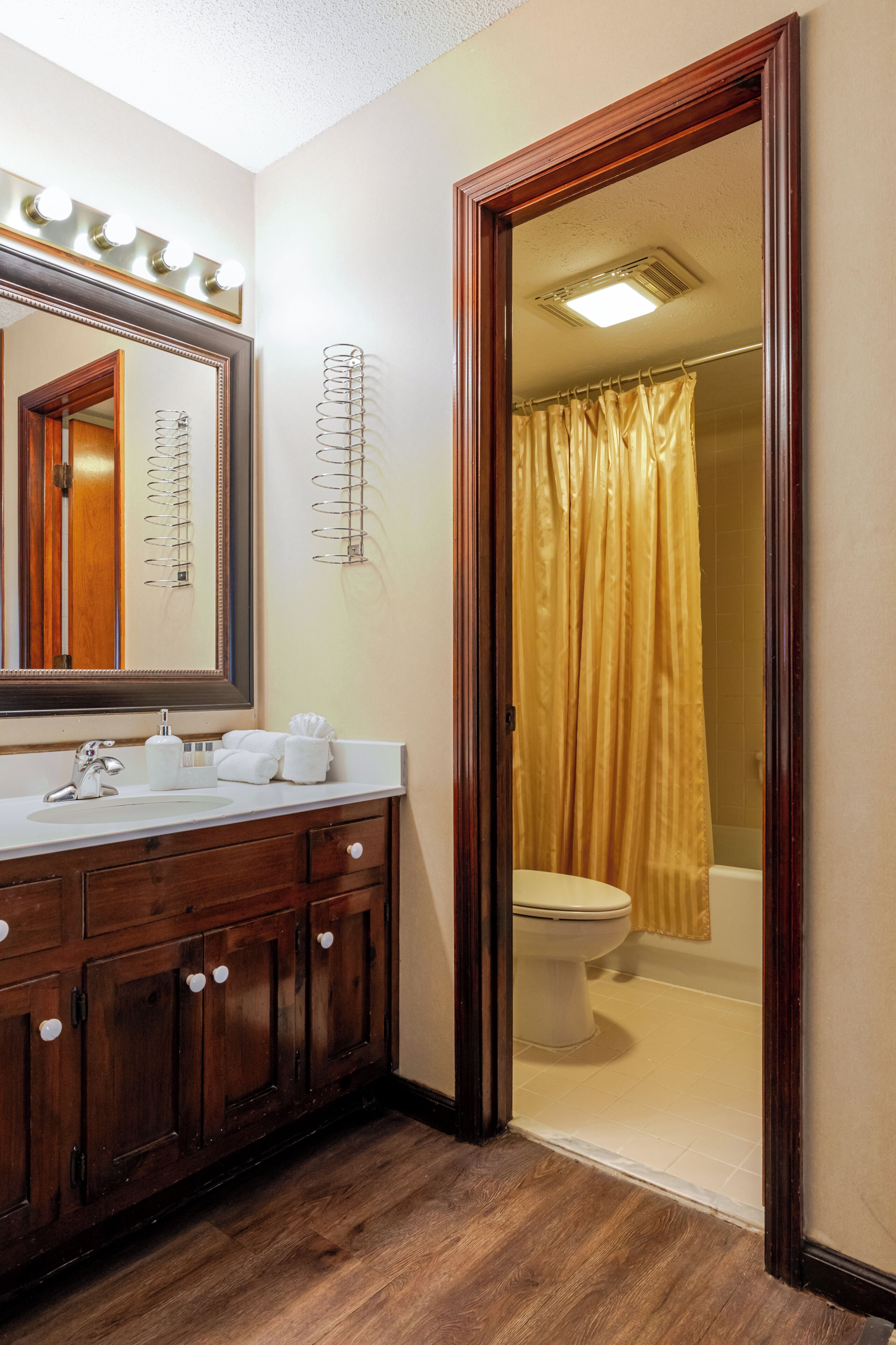 A bathroom with a wooden vanity, a large mirror with lights, a toilet, and a shower curtain.