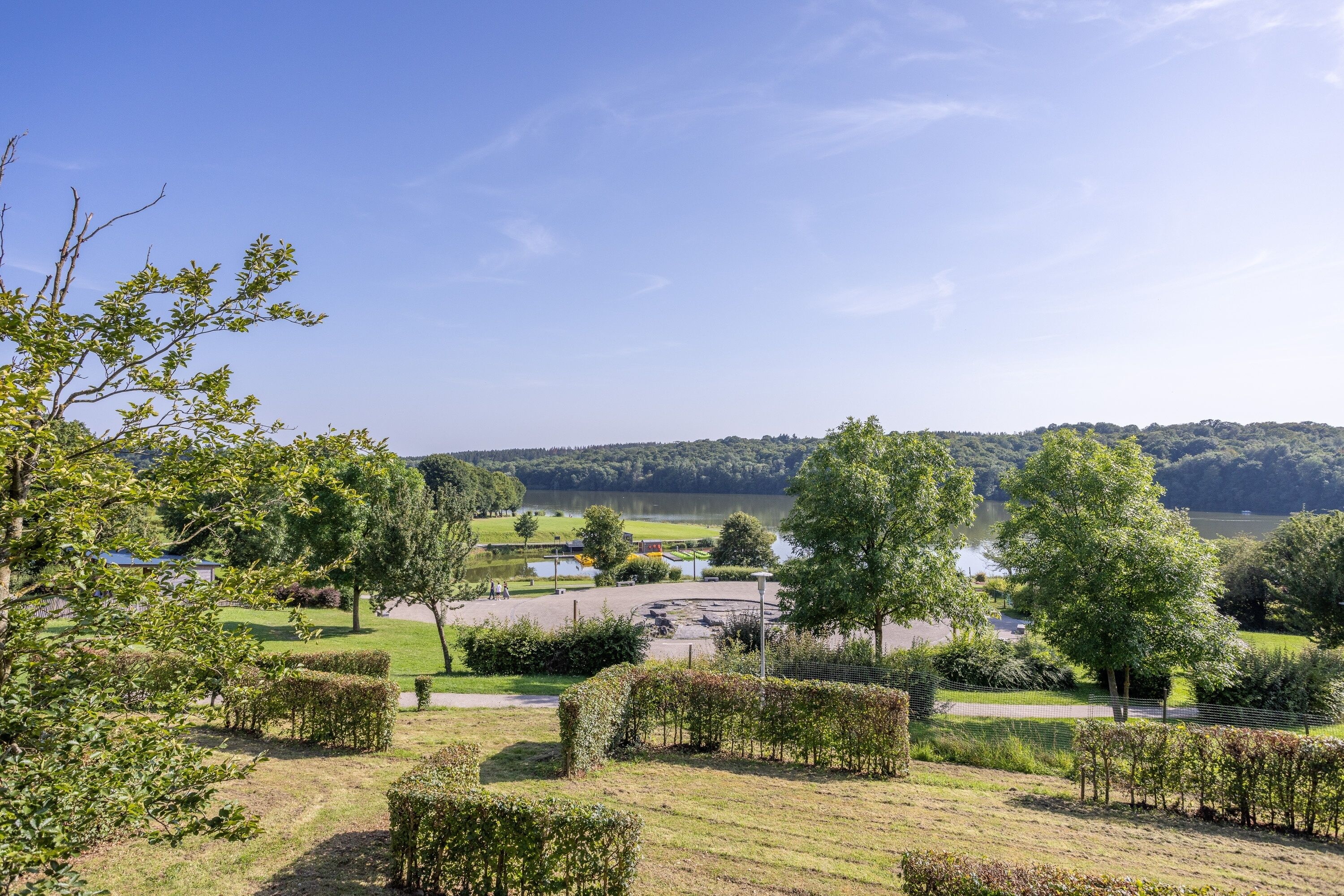 Een aangelegde tuin met strakke heggen, een rond pad en een waterpartij op de achtergrond.