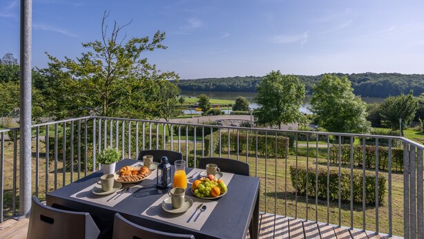Een balkon met een tafel, ontbijtartikelen, uitzicht op een meer en omliggend groen.