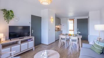 Uma sala de estar moderna com televisão de tela plana, uma área de jantar com mesa e cadeiras e uma cozinha ao fundo.