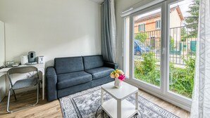 Un salon moderne avec un canapé gris, une petite table blanche ornée d’un vase de fleurs, une chaise en métal et une grande fenêtre donnant sur un jardin verdoyant et des bâtiments.