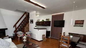 Un vaste salon avec un canapé blanc, une table basse en bois et un coin repas comprenant une table et des chaises. On aperçoit au fond une cuisine équipée de placards et d’un téléviseur.