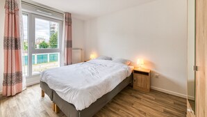 A bedroom with a bed, bedside lamps, a wooden floor, and a view through a large window. - Moulin à Vent A14 - Appt pour 4 avec balcon (Bondy)