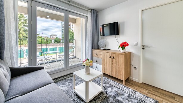A modern living room with a sofa, a small white table with a vase of flowers, a wooden cabinet, and a TV mounted on the wall. - Moulin à Vent A14 - Appt pour 4 avec balcon (Bondy)