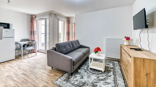 A modern living room with a grey sofa, a small white table with a vase of flowers, a wooden sideboard, and a mounted TV. - Moulin à Vent A21 - Appt pour 4 (Bondy)
