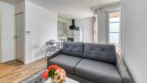 A modern living room with a grey sofa, a dining table with chairs, a kitchen area, and a view of the outdoors through a window. - Moulin à Vent A21 - Appt pour 4 (Bondy)