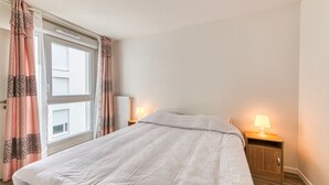 A bedroom with a bed, a nightstand with a lamp, a window with curtains, and a radiator. - Moulin à Vent A21 - Appt pour 4 (Bondy)
