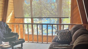 A cozy room with a wooden ceiling, a ceiling fan, a cushioned bench, a sofa with patterned cushions, and a large window with a view of trees.