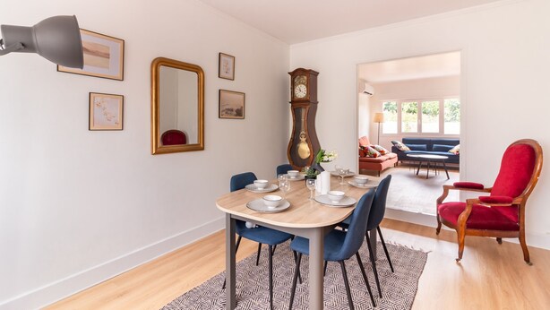 A dining area with a wooden table set for four, blue chairs, a red chair, and a large wall clock.
