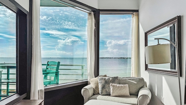 Una habitación con sofá, reposapiés y una mesa, ventanas amplias con vista al mar y una isla distante.