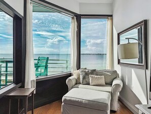Una habitación con sofá, reposapiés y una mesa, ventanas amplias con vista al mar y una isla distante.