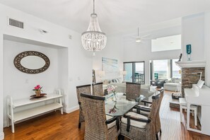 A spacious living area with a dining table, wicker chairs, a chandelier, a piano, and a fireplace.
