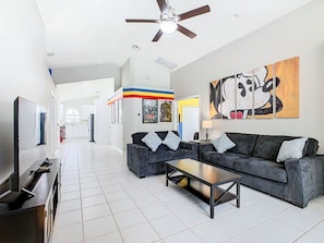 A modern living room with a sofa, coffee table, and TV. There is a ceiling fan, artwork on the wall, and a hallway leading to other rooms. - Disney Themed 5BR Villa w/ Pool & Spa + Game Room (Kissimmee)