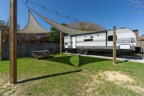 A recreational vehicle (RV) with a "SPORTSMEN" decal parked in a backyard with a picnic table, a shade sail, and a wooden fence. - Coleman Camper Oasis-Beaches-shopping-dining (Pensacola)