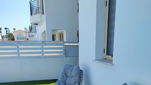 Dos sillas de mimbre grises con cojines sobre un césped artificial verde, una pequeña mesa negra y un edificio blanco con balcones al fondo.