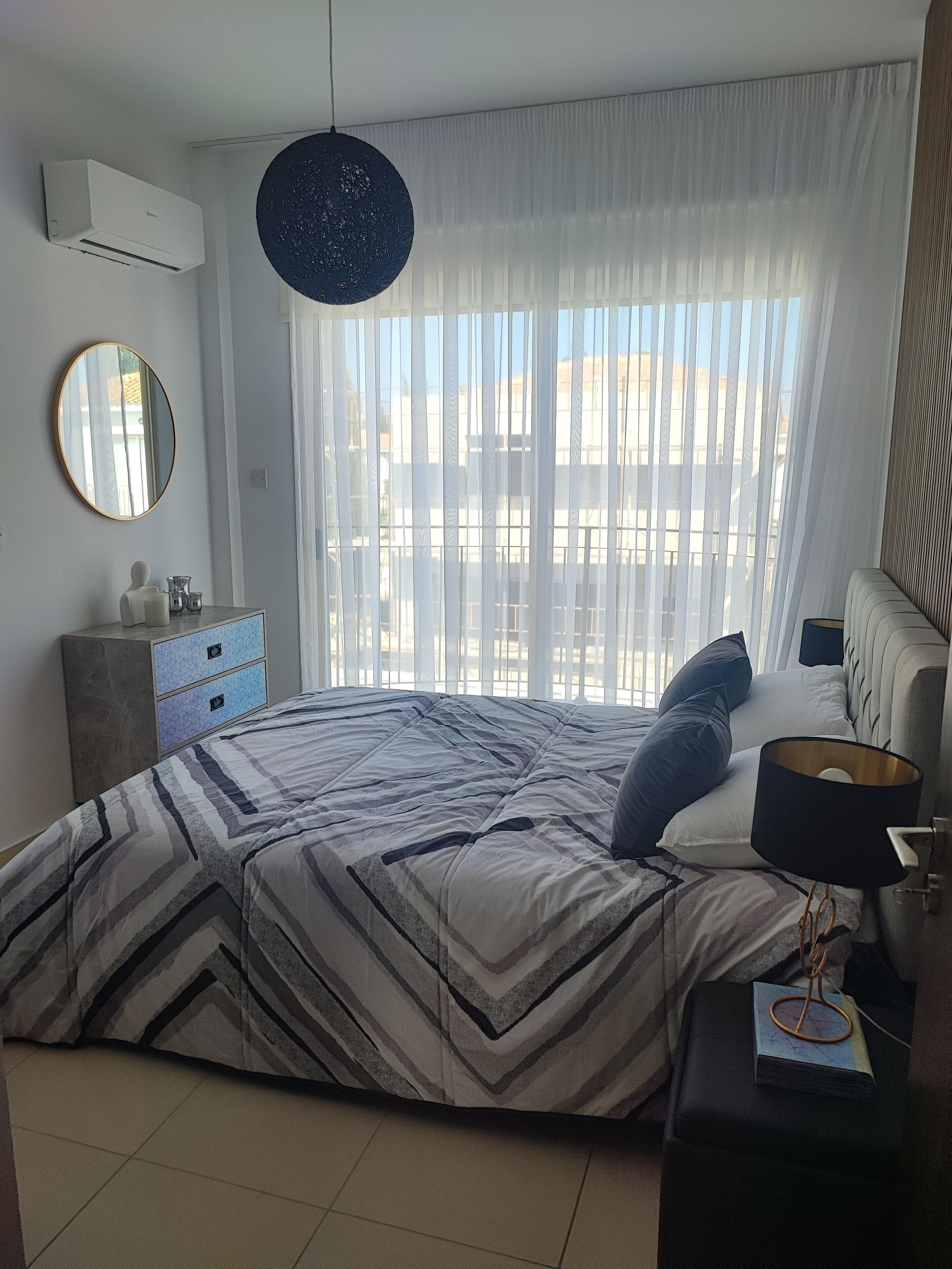 Een slaapkamer met een bed, nachtkastje, lamp, dressoir, spiegel en een raam met dunne gordijnen.