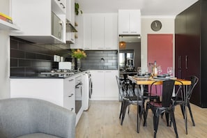 A modern kitchen with white cabinetry, a dining area with a wooden table and black chairs, a red door, and a clock on the wall. - Cape on Porterfield 5 (Cape Town)