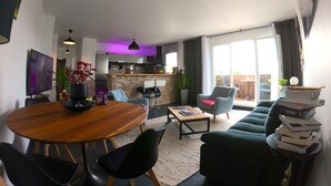 A modern living room with a wooden dining table, black chairs, a green sofa, and a coffee table. There is a kitchen in the background with a purple light, and a balcony with a view. - Elegant 70m2 Rooftop at the foot of Le Bourget station (Le Bourget)