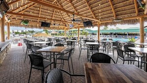 Un espace extérieur aménagé en terrasse, avec des tables et des chaises, un toit de chaume et une vue sur une piscine et la verdure.