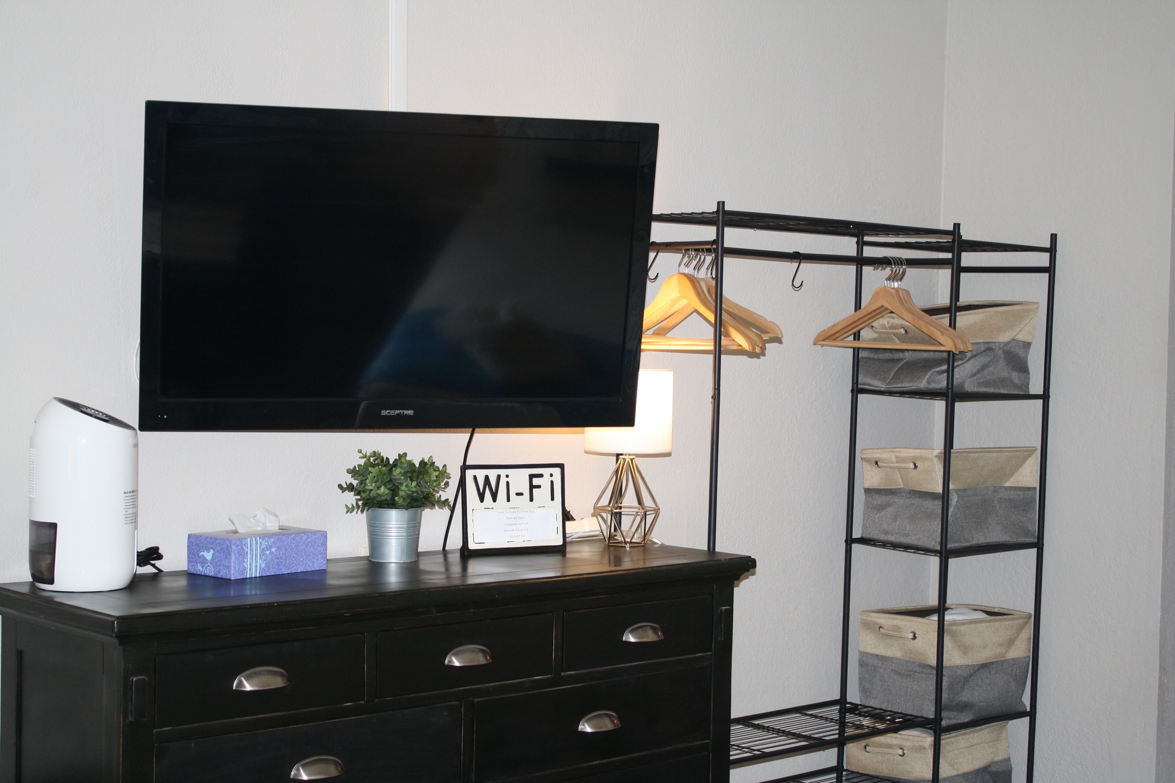 A flat-screen TV mounted on a dresser, a Wi-Fi sign, a lamp, and a storage rack with hanging clothes and storage boxes.