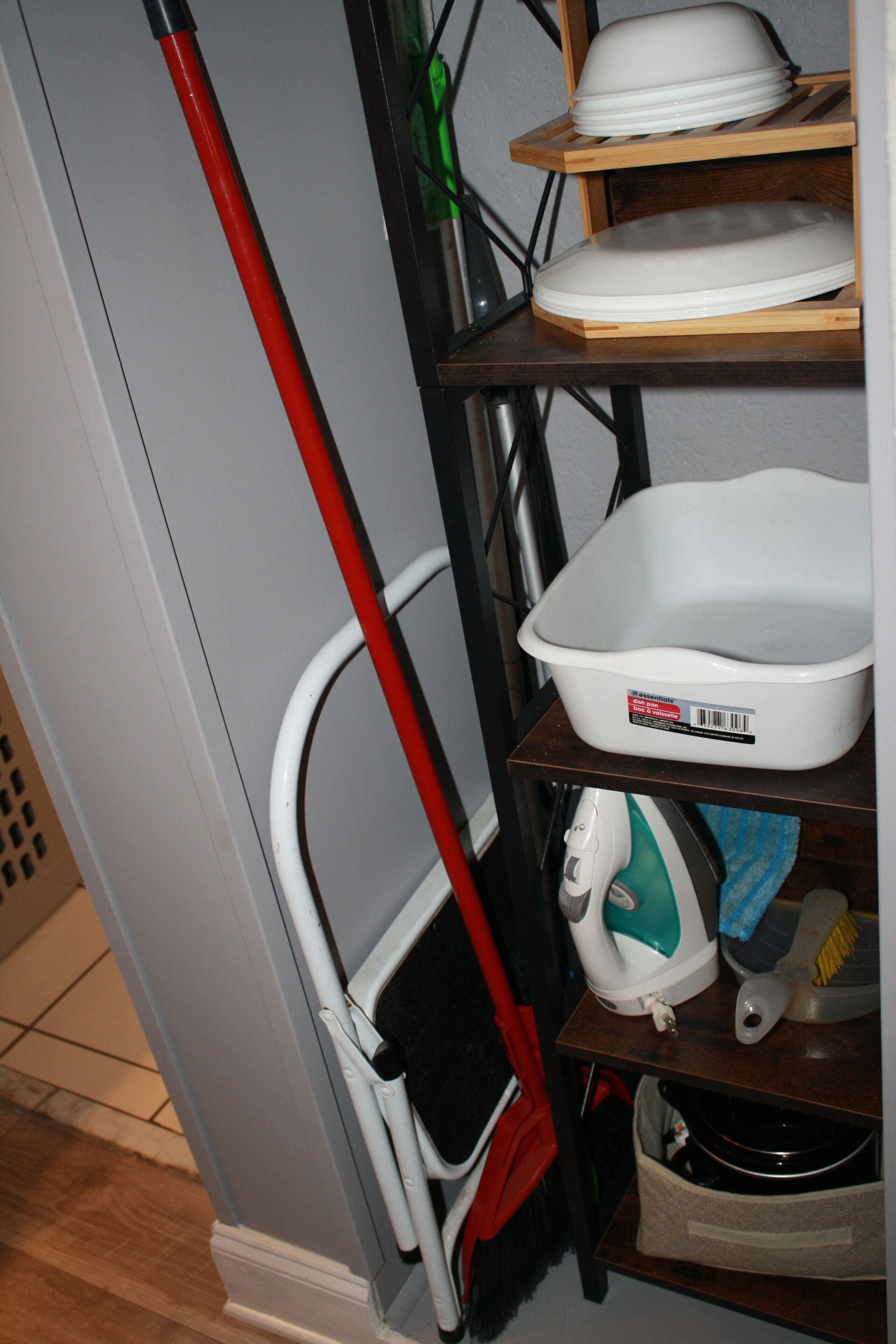 A corner of a room with a mop, a white basin, a dish rack, and a shelf with a white appliance.