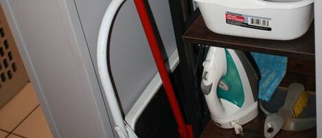 A corner of a room with a mop, a white basin, a dish rack, and a shelf with a white appliance.