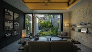 A modern living room with a sofa, chairs, a coffee table, and a lamp. There is a large window with a view of a garden and a stone wall.
