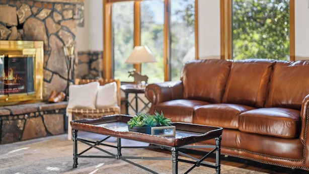 A living room with a leather sofa, a coffee table with a welcome sign, a fireplace, and a view of greenery outside. - Country Club (Flagstaff)
