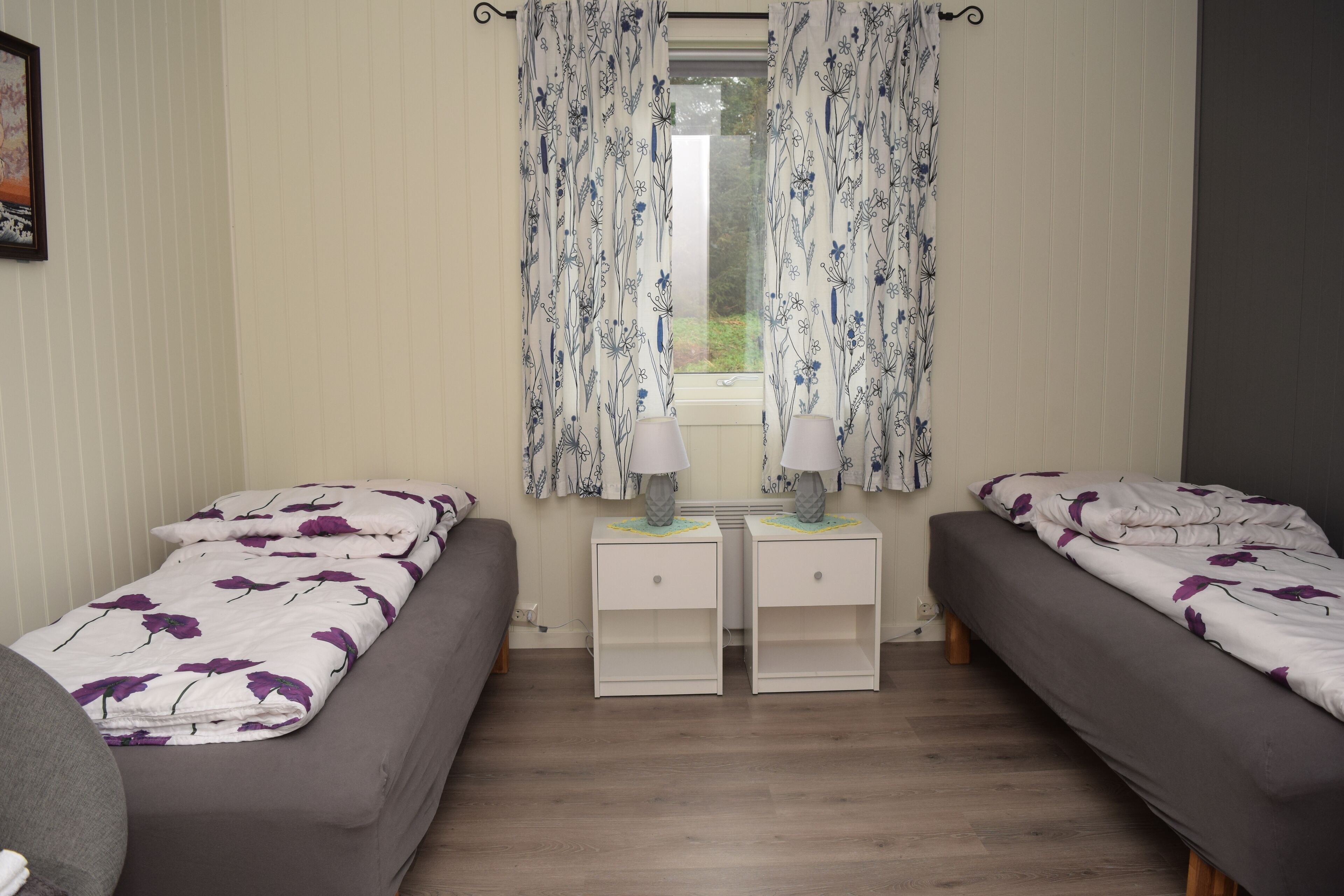 A bedroom with two single beds, a window with floral curtains, bedside tables with lamps, and a grey sofa.