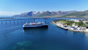 A ferry named "HAVILA KYSTUTEN" crossing a bridge over a body of water with a mountainous landscape in the background.