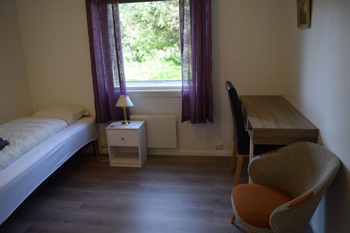 A bedroom with a bed, a nightstand with a lamp, a chair, and a desk.