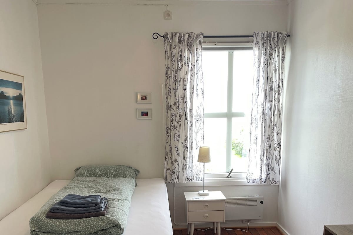 A bedroom with a bed, a nightstand, a window with curtains, a radiator, and a small table.