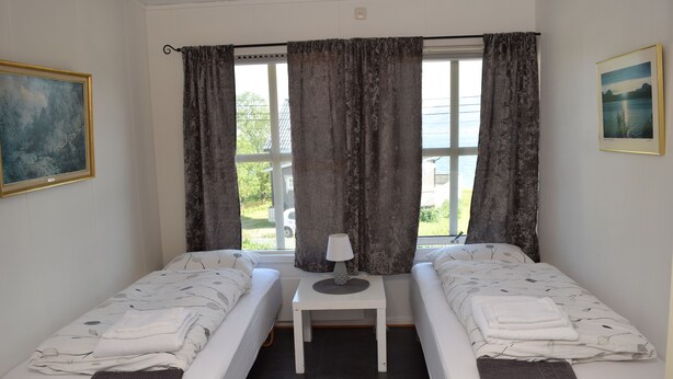 A room with two single beds, a small table with a lamp, a window with curtains, and a painting on the wall.