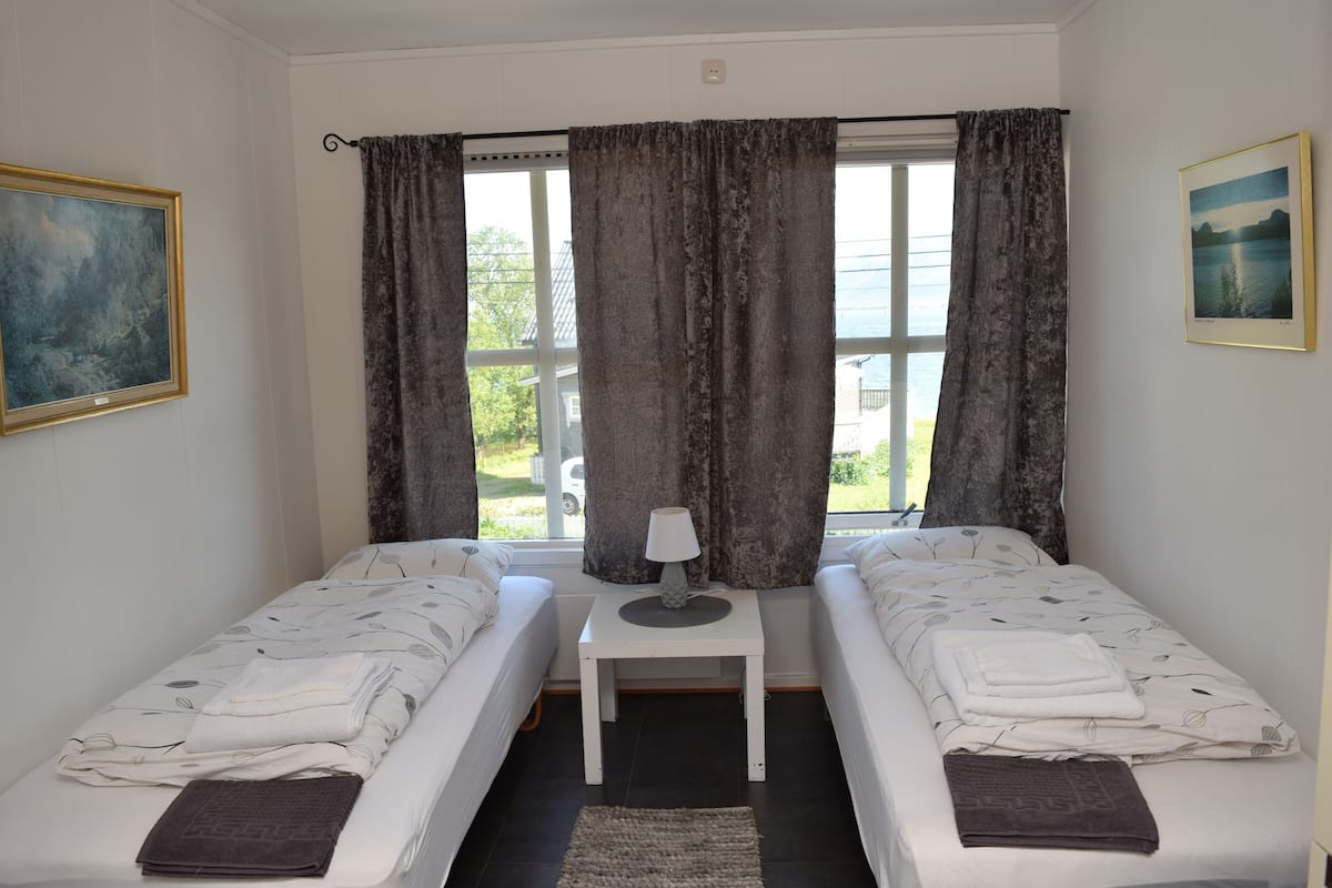 A room with two single beds, a small table with a lamp, a window with curtains, and a painting on the wall.