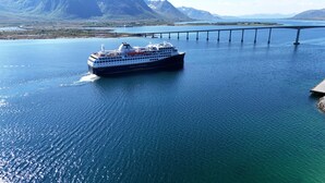 A large cruise ship named 'Havila Kystoten' sailing on a calm body of water with a bridge in the background.