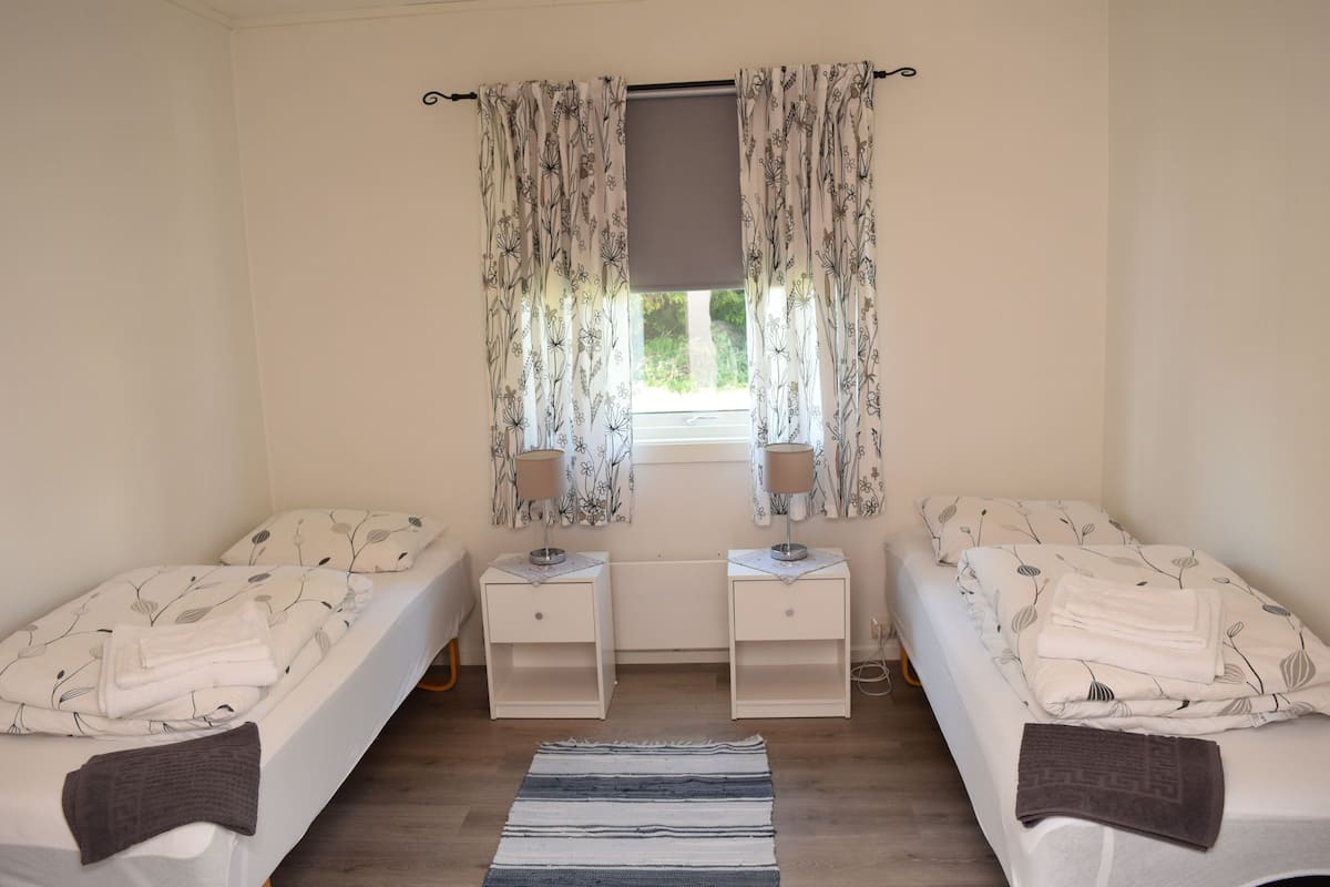 A twin bedroom with two beds, a window with curtains, bedside tables with lamps, and a striped rug.