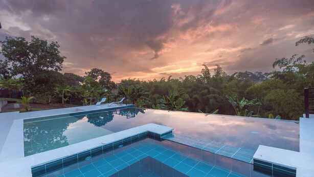 A rectangular infinity pool with clear blue tiles, surrounded by lush greenery and trees, reflecting the sky.