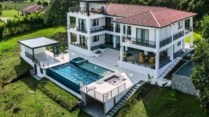 A modern two-story house with a swimming pool, surrounded by lush greenery and a clear sky.