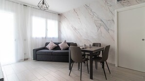 A modern living room with a dark grey sofa, two patterned pillows, a dining table with four chairs, and a marble-patterned wall. - Italianway Comfort - Fara 29 (Milan)