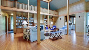 A spacious loft-style room with a high ceiling, wooden floors, and a mezzanine level with bookshelves.