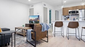 Una sala moderna con un televisor de pantalla plana, un sofá, una mesa de centro y una zona de comedor con taburetes de bar.